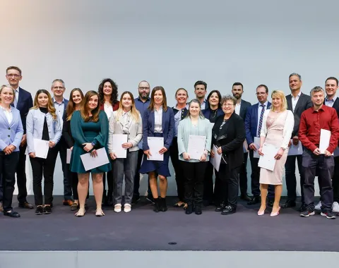 Eine Gruppe von Absolventinnen und Absolventen steht auf einer Bühne und hält Zertifikate in den Händen. Sie lächeln in die Kamera. Die Personen sind formell gekleidet, und im Hintergrund ist eine neutrale Wand zu sehen.