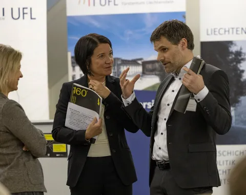 Drei Personen stehen in einem Gespräch vor Roll-Up-Bannern der Universität Liechtenstein und des Liechtenstein-Instituts. Eine Person hält ein Buch in der Hand. Die Szene findet in einem formellen Rahmen statt, vermutlich bei einer akademischen Veranstaltung.