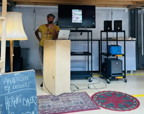 Ein Mann steht an einem Rednerpult in einem Raum mit Holzdach. Neben ihm ist ein Bildschirm mit einer Präsentation und ein Schild mit der Aufschrift „Architecture by Dessert, Herwig Bauer, Poolbarfestival“. Der Raum ist mit Teppichen und Stehlampen dekoriert.