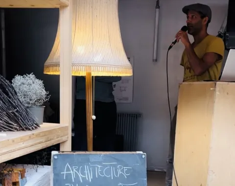 Ein Mann spricht in ein Mikrofon neben einem Rednerpult. Im Vordergrund steht eine Tafel mit der Aufschrift „Architecture by Dessert, Herwig Bauer, Poolbarfestival“. Links sind ein Regal und eine Stehlampe zu sehen.