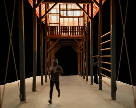 Ein Modell eines architektonischen Entwurfs zeigt eine überdachte Struktur mit sichtbaren Holzbalken. Eine Person läuft auf einem Weg unter der Konstruktion. Das Design kombiniert traditionelle Bauweise mit modernen Elementen und betont lineare Formen.