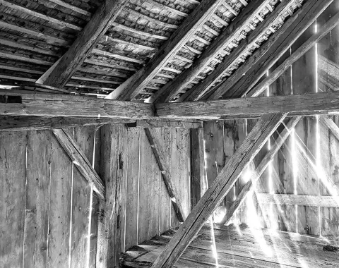Schwarz-Weiß-Foto eines alten Dachbodens mit sichtbaren Holzbalken und einfallendem Licht durch die Dachlücken. Die Struktur zeigt traditionelle Bauweise und vermittelt eine rustikale Atmosphäre.