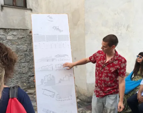 Ein Student präsentiert architektonische Skizzen auf einem großen Plakat in einem historischen Dorf. Um ihn herum stehen und sitzen mehrere Personen, die aufmerksam zuhören. Die Szene spielt sich in einem gepflasterten Außenbereich ab.