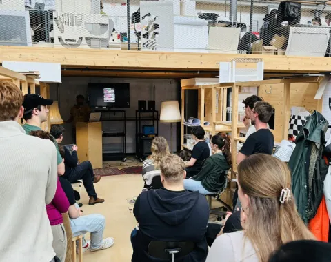 Eine Gruppe von Studierenden sitzt in einem Architekturatelier und hört einem Vortrag zu. Im Hintergrund sind Regale und Arbeitsmaterialien zu sehen.