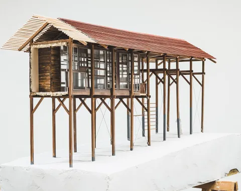 Ein Architekturmodell eines Strickbaus auf Stelzen, das auf einem weißen Sockel präsentiert wird. Das Modell zeigt eine lineare Struktur mit einem Satteldach und großen Fenstern, die traditionelle Bauweise und moderne Elemente kombinieren.