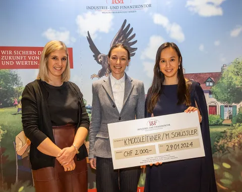 Drei Personen stehen lächelnd vor einem Hintergrund mit dem Logo der Industrie- und Finanzkontor. Eine Person hält einen großen Scheck mit der Aufschrift "CHF 2,000" und einem Datum. Im Hintergrund ist ein stilisiertes Bild mit einem Adler und einem Haus zu sehen.