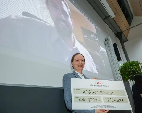 Eine Person hält einen großen Scheck mit der Aufschrift "Adrian Vogler, CHF 4,000, 29.01.2024" in einem Vortragsraum. Im Hintergrund ist eine Projektion eines lächelnden Mannes auf einer Leinwand zu sehen.