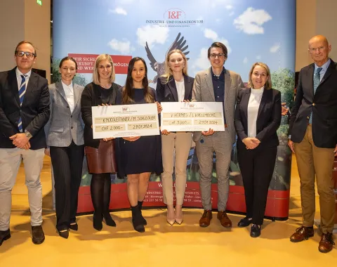 Gruppe von acht Personen posiert mit zwei großen Schecks bei der Verleihung des I&F Family Wealth Preservation Awards an der Universität Liechtenstein. Die Studierenden wurden für ihre herausragenden Strukturierungsvorschläge ausgezeichnet. Im Hintergrund ist ein Banner des Industrie- und Finanzkontors zu sehen.