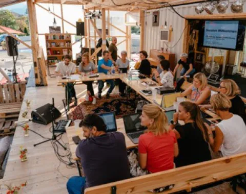 Eine Gruppe von Menschen sitzt in einem offenen, modularen Raum an einem Tisch mit Laptops. Der Raum ist aus Holz und enthält Bücherregale und technische Ausrüstung. Es herrscht eine lebhafte Diskussionsatmosphäre.