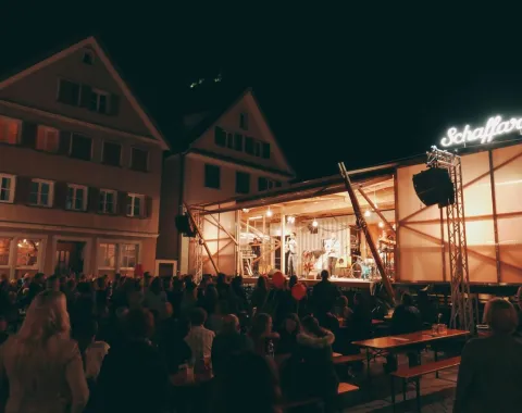 Eine beleuchtete Bühne des Base Camps der Universität Liechtenstein bei Nacht, umgeben von einem Publikum. Die Bühne ist modular und mobil, passend zur temporären Nutzung für Veranstaltungen und Stadtentwicklungsprojekte.