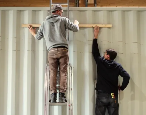 Drei Personen arbeiten an einem großen, weißen Container. Zwei stehen auf Leitern und befestigen etwas an der Wand, während die dritte Person unten steht und assistiert.