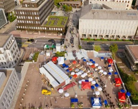 Luftaufnahme eines städtischen Platzes mit dem Base Camp der Universität Liechtenstein. Zu sehen sind modulare Strukturen, bunte Zelte und Menschen, die sich auf dem Platz bewegen. Gebäude umgeben den Platz, und eine Straße verläuft im Hintergrund.