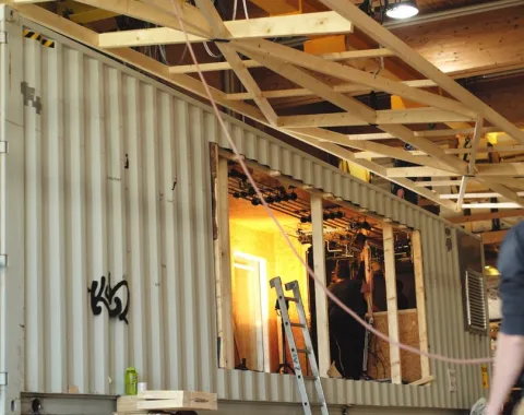 Ein großer, umgebauter Container in einer Werkhalle, mit sichtbaren Holzverstärkungen und einer Leiter, symbolisiert die flexible Nutzung des Base Camps der Universität Liechtenstein für Stadtentwicklungsprojekte.