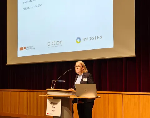 Eine Frau steht an einem Rednerpult vor einer Leinwand mit der Aufschrift „Herzlich willkommen! Compliance-Day 2024, Universität Liechtenstein, Schaan, 14. Mai 2024“. Im Hintergrund sind Logos von Sponsoren zu sehen.