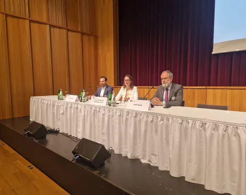 Podiumsdiskussion beim Compliance-Day 2024 an der Universität Liechtenstein. Drei Personen sitzen hinter einem Tisch mit Mikrofonen und Getränken. Der Raum hat Holzwände und einen roten Vorhang im Hintergrund.