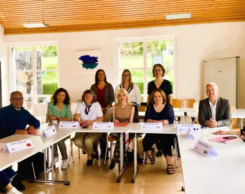 Eine Gruppe von zwölf Personen sitzt an einem U-förmigen Tisch in einem hellen Raum. Sie sind Teil eines internationalen Besuchsprogramms an der Universität Liechtenstein. Im Hintergrund sind große Fenster und eine Holzbalkendecke zu sehen.