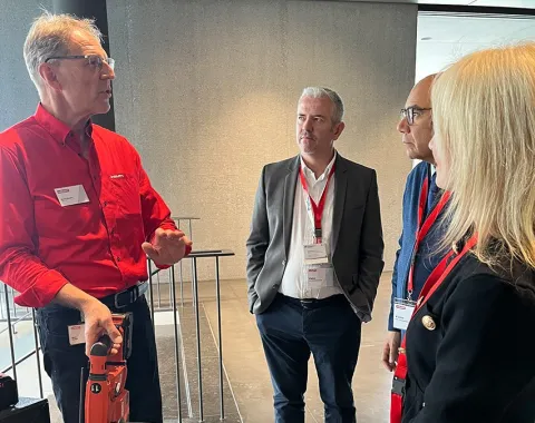 Eine Gruppe von vier Personen steht in einem modernen Gebäude. Eine Person hält ein Werkzeug und erklärt etwas den anderen. Alle tragen Namensschilder und hören aufmerksam zu.