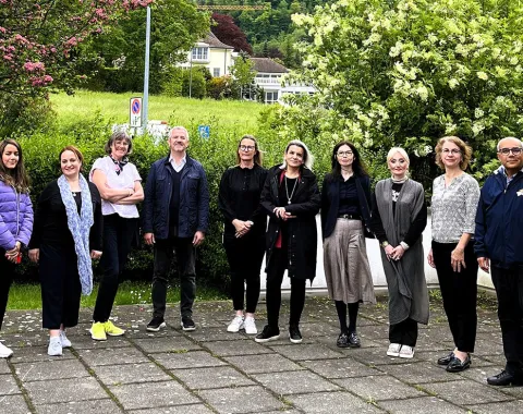 Eine Gruppe von zehn Personen steht draußen auf einem gepflasterten Weg vor einer grünen Hecke und blühenden Bäumen. Die Personen sind in Freizeitkleidung und lächeln in die Kamera. Im Hintergrund sind ein Gebäude und eine Wiese zu sehen.