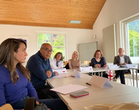 Eine Gruppe von Personen sitzt an einem Konferenztisch in einem hellen Raum mit großen Fenstern. Sie diskutieren angeregt, vor ihnen liegen Notizblöcke und Namensschilder. Im Hintergrund sind grüne Bäume und Berge zu sehen.