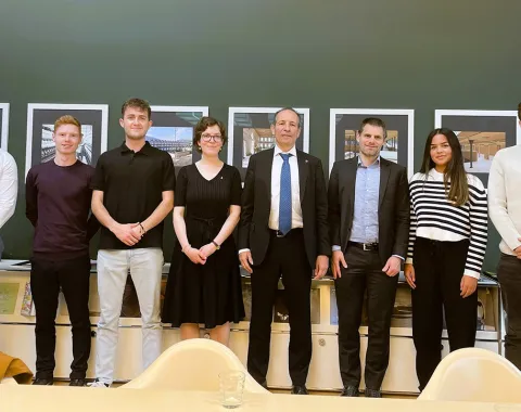 Eine Gruppe von sieben Personen, darunter Studierende und zwei Männer in Anzügen, steht lächelnd vor einer Wand mit Bildern. Sie befinden sich in einem Raum der Universität Liechtenstein.