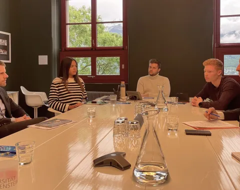 Eine Gruppe von Personen sitzt an einem Konferenztisch in einem Raum mit großen Fenstern. Sie diskutieren in einer formellen Umgebung. Auf dem Tisch stehen Wassergläser und Karaffen. Im Hintergrund sind grüne Bäume und ein bewölkter Himmel zu sehen.