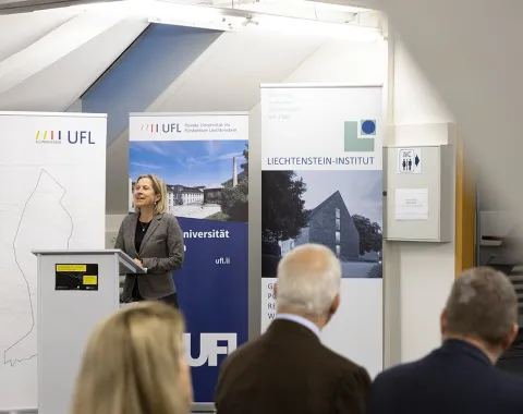 Eine Frau steht an einem Rednerpult vor Bannern der Universität Liechtenstein, der UFL und des Liechtenstein-Instituts. Im Vordergrund sind unscharf einige Zuhörer zu sehen.
