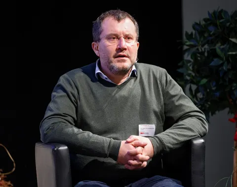 Eine Person sitzt auf einem schwarzen Sessel und hält die Hände gefaltet. Im Hintergrund sind Pflanzen zu sehen.