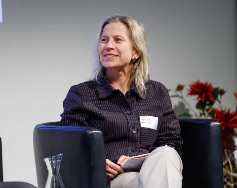 Eine Person sitzt lächelnd auf einem Podium in einem Konferenzraum. Im Hintergrund sind Blumenarrangements zu sehen.