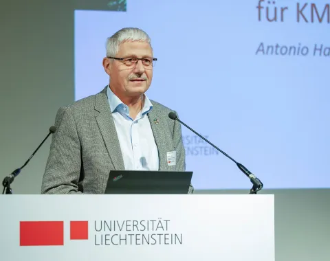 Ein Redner steht an einem Podium mit dem Logo der Universität Liechtenstein und spricht in ein Mikrofon. Im Hintergrund ist eine Präsentationsfolie zu sehen.