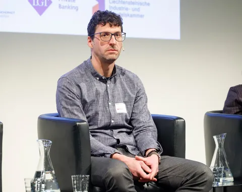 Eine Person sitzt auf einer Bühne in einem schwarzen Sessel. Im Hintergrund sind Logos von Sponsoren oder Partnern zu sehen. Auf dem Tisch vor der Person stehen Wasserkaraffen und Gläser.