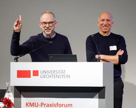 Zwei Männer stehen lächelnd hinter einem Rednerpult mit dem Logo der Universität Liechtenstein. Einer hebt die Hand, während der andere die Arme verschränkt. Im Vordergrund sind rote Blumen zu sehen.