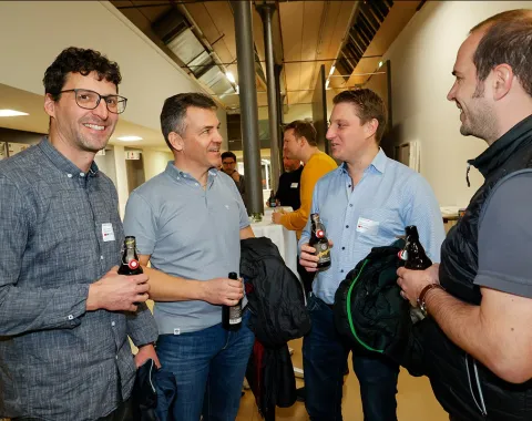 Vier Männer unterhalten sich bei einer Veranstaltung an der Universität Liechtenstein. Sie halten Getränke in der Hand und tragen Namensschilder. Die Atmosphäre wirkt entspannt und gesellig.
