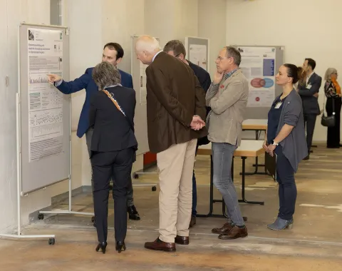 Eine Gruppe von Personen betrachtet eine wissenschaftliche Posterausstellung in einem Innenraum. Ein Mann zeigt auf ein Poster und erklärt den Inhalt. Im Hintergrund sind weitere Poster und Personen zu sehen.