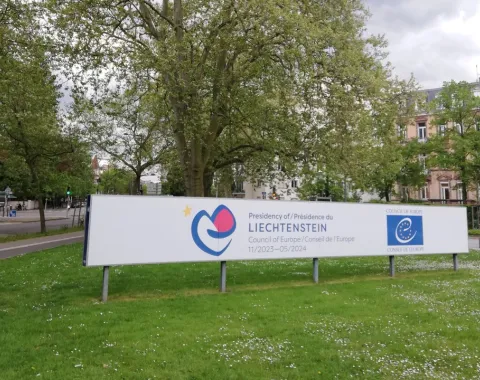 Ein großes Schild mit dem Logo des Fürstentums Liechtenstein und dem Europarat steht auf einer grünen Wiese, umgeben von Bäumen und Gebäuden im Hintergrund.