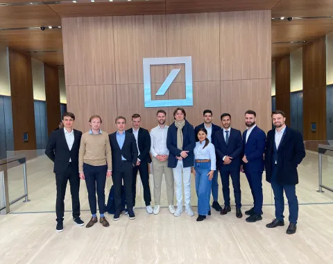 Eine Gruppe von Studierenden und Begleitpersonen steht in einem modernen Foyer vor dem Logo einer bekannten Bank. Sie tragen formelle Kleidung und lächeln in die Kamera.