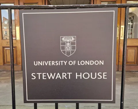 Ein Schild vor dem Eingang eines Gebäudes mit der Aufschrift "University of London, Stewart House". Im Hintergrund sind Holztüren und die Hausnummer 32 zu sehen.