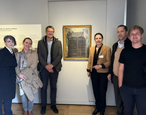 Eine Gruppe von sechs Personen steht in einem Ausstellungsraum vor einem Gemälde. Sie lächeln in die Kamera. Der Raum hat helle Wände und einen Holzboden.