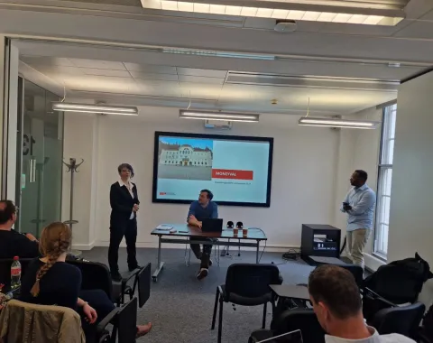 Ein Vortrag im Seminarraum: Eine Person steht links und spricht, während eine andere Person rechts steht. In der Mitte sitzt jemand an einem Tisch. Im Hintergrund ist eine Präsentation auf einem Bildschirm zu sehen. Zuschauer sitzen im Vordergrund.