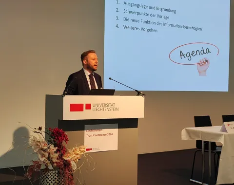 Ein Redner steht an einem Pult mit dem Logo der Universität Liechtenstein. Im Hintergrund ist eine Präsentationsfolie mit der Überschrift "Agenda" zu sehen. Links neben dem Pult steht eine Blumendekoration.