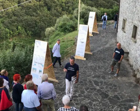 Eine Gruppe von Menschen betrachtet im Freien aufgestellte Tafeln mit Informationen. Die Szene spielt sich auf einem gepflasterten Weg neben einem alten Steingebäude ab, umgeben von üppiger, grüner Landschaft.