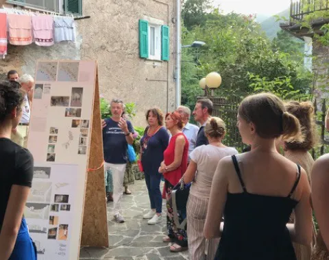 Eine Gruppe von Menschen betrachtet eine Ausstellung im Freien in einem historischen Dorf. Im Vordergrund sind Informationstafeln zu sehen, im Hintergrund eine steinerne Hausfassade und grüne Hügel.