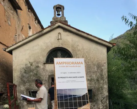 Ein Mann steht vor einem historischen Gebäude in Castè, Italien, und betrachtet ein Plakat mit der Aufschrift "Amphiorama". Das Plakat informiert über ein Projekt der Universität Liechtenstein im Rahmen des Advanced Studios Upcycling. Im Hintergrund sind die malerischen Hügel der Cinque Terre zu sehen.