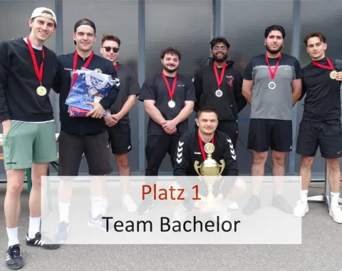 Acht junge Männer stehen in sportlicher Kleidung vor einer Wand. Sie tragen Medaillen um den Hals. Einer kniet vorne und hält einen Pokal. Text im Bild: "Platz 1 Team Bachelor".