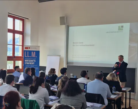 Ein Vortragender steht vor einer Gruppe von Teilnehmenden in einem Seminarraum der Universität Liechtenstein. Auf der Leinwand ist eine Präsentationsfolie mit dem Text "Besten Dank für Ihre Aufmerksamkeit!" zu sehen. Im Hintergrund ist ein Banner mit der Aufschrift "LL.M. Wirtschafts-Strafrecht" sichtbar.