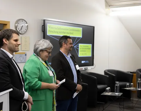 Drei Personen stehen nebeneinander in einem Konferenzraum. Im Hintergrund ist ein Bildschirm mit der Aufschrift "Wissenschaftsverbund Liechtenstein" zu sehen. Links im Bild ist ein Rednerpult, rechts sind leere Stühle und Tische. Die Personen tragen Namensschilder und lächeln.