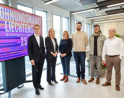 Gruppe von sechs Personen steht vor einem Bildschirm mit der Aufschrift "Wohnungsmarkt Liechtenstein". Die Personen lächeln und sind in einem modernen Raum mit großen Fenstern.