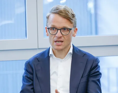 Ein Mann in einem Anzug spricht gestikulierend während einer Podiumsdiskussion in einem modernen Raum mit großen Fenstern.