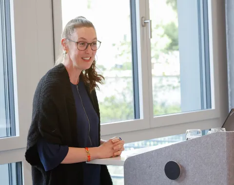 Eine Person steht lächelnd an einem Rednerpult in einem modernen Raum mit großen Fenstern. Sie trägt eine Brille und hält einen Stift in der Hand. Im Hintergrund sind Bäume zu sehen.