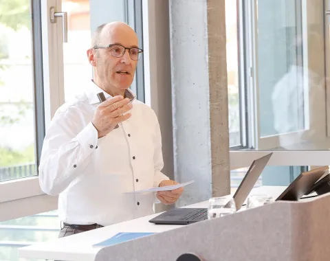 Ein Mann hält einen Vortrag in einem modernen Raum mit großen Fenstern. Er steht an einem Rednerpult mit Laptop und Notizen.