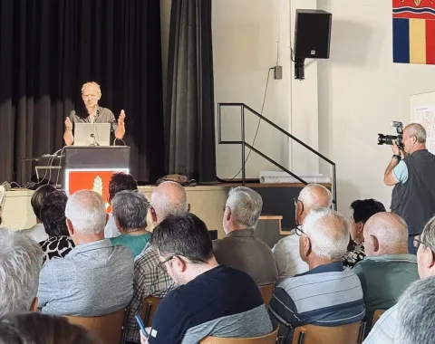 Ein Vortragender steht an einem Rednerpult vor einem Publikum in einem Saal. Im Hintergrund sind schwarze Vorhänge und ein Fotograf macht Bilder. An der Wand hängt ein Wappen.